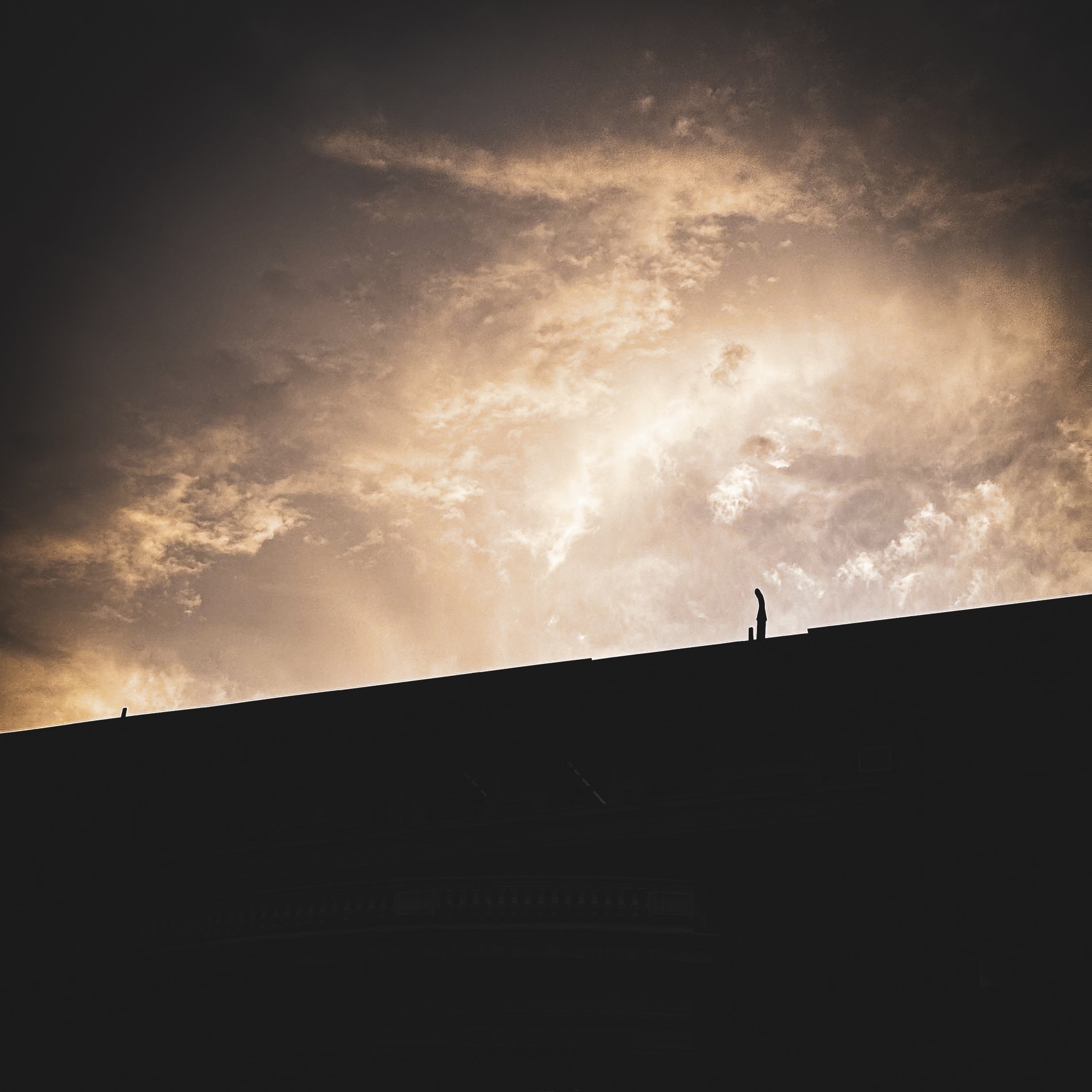 A lone figure silhouetted on a steep roofline against a burning cloud sky