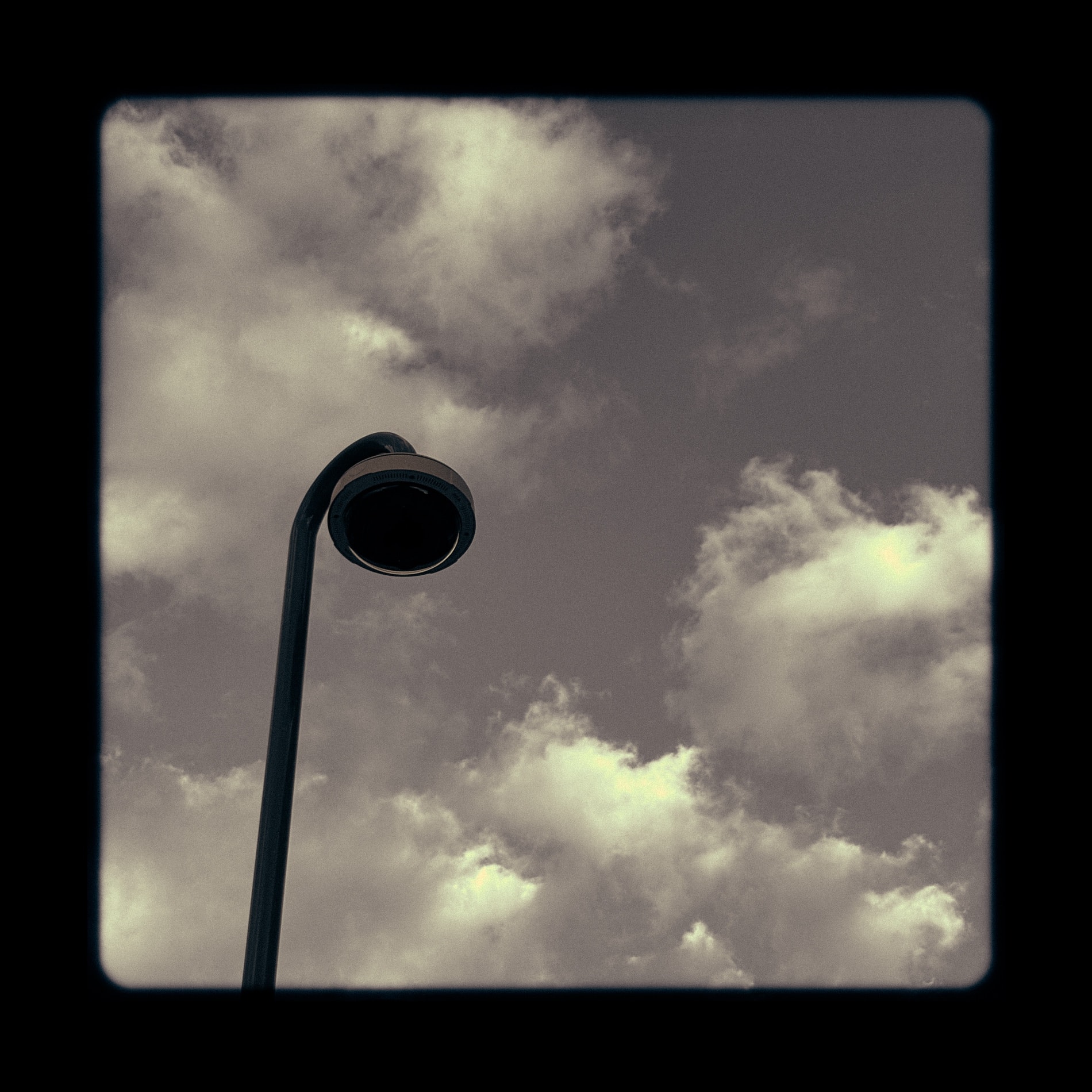 A streetlamp head against banked cloud, framed in a soft black vignette